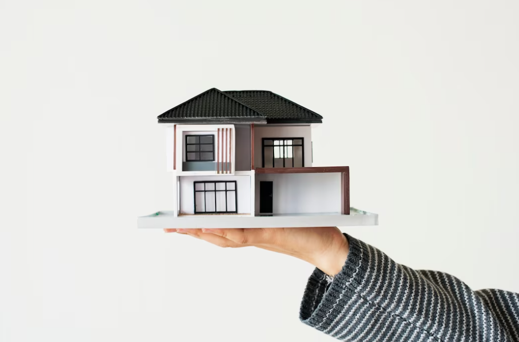 a person holds a small model of a house with a black roof in front of a white wall