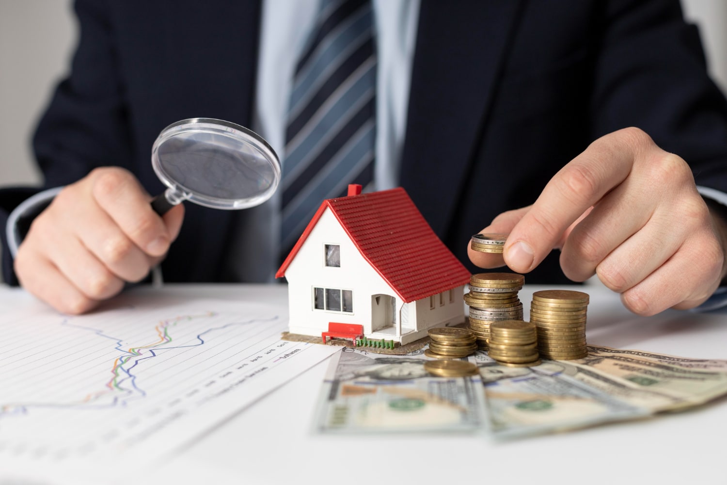 a man holds a magnifying glass over a toy house on a table, with banknotes and stacks of coins