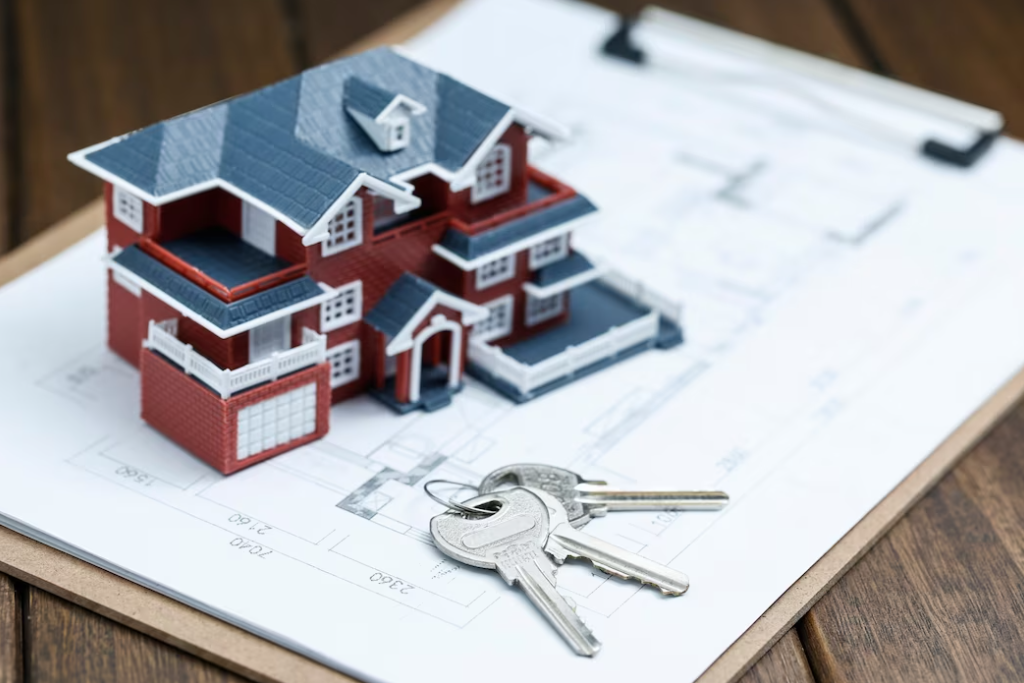 a small model of a red house with a black roof, the keys, and sheets with house planning on the dark wooden table